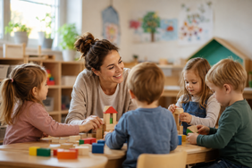 Eine Erzieherin sitzt mit vier Kindern in einem hellen Gruppenraum an einem Tisch und baut gemeinsam mit ihnen aus bunten Holzklötzen. Im Hintergrund sind Regale, Pflanzen und Kinderbilder zu sehen.