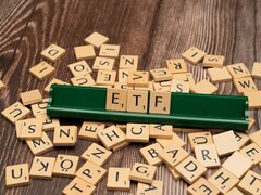 Eine Vielzahl von Scrabble-Buchstaben liegt ungeordnet auf einem dunklen Holztisch. Im Zentrum steht ein grünes Buchstaben-Bänkchen, auf dem die Buchstaben „E, T, F“ ordentlich aufgereiht sind. Das Bild symbolisiert das Thema Geldanlage in börsengehandelte Indexfonds.