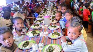 In einer hellen Schulkantine in Tsarahonenana sitzen zahlreiche Kinder an langen Tischen, die mit bunt gestreiften Decken bedeckt sind, und essen eine Mahlzeit aus Reis und grünem Gemüse.