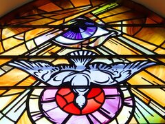 Ein farbenprächtiges Kirchenfenster aus Glasmalerei zeigt im oberen Teil ein wachsames Auge und darunter eine herabfliegende Taube vor einem strahlenförmigen Hintergrund.