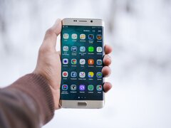 Eine Hand hält ein Smartphone vor einem unscharfen, hellen Hintergrund. Auf dem Display ist das App-Menü mit zahlreichen Icons wie Google Chrome, Facebook und Instagram zu sehen.