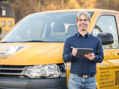 Person mit Tablet steht vor einem gelben KAB‑Transporter, der im Freien geparkt ist.