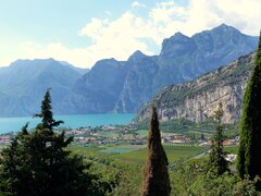 Panoramablick auf den Gardasee in Italien: Türkisfarbenes Wasser glitzert vor einer imposanten Kulisse aus steilen, felsigen Bergen. Im Vordergrund erstreckt sich eine grüne Ebene mit Weinreben und Zypressen, die das mediterrane Flair unterstreichen.