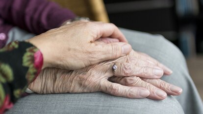 Die Hand einer jüngeren Person liegt schützend und stützend auf der deutlich faltigeren Hand einer älteren Person, die einen Ring trägt.