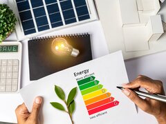 Draufsicht auf einen Schreibtisch mit Utensilien zur Energieeffizienz: Eine Hand hält eine Energie-Skala (A-G) und einen Zweig. Drumherum liegen ein Taschenrechner mit der Anzeige „SAVE“, eine leuchtende Glühbirne, ein Solarpanel und ein Hausmodell.