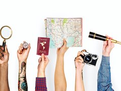 Sechs verschiedene Arme ragen von unten ins Bild und halten diverse Gegenstände in die Höhe, die das Thema Reisen und Entdecken symbolisieren: eine Lupe, einen Kompass, einen Reisepass, eine Landkarte, eine Kamera und ein Fernrohr.
