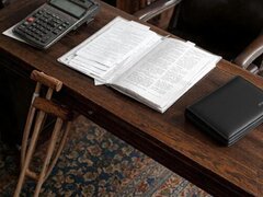 Holzschreibtisch mit Taschenrechner, geöffnetem Buch und schwarzer Agenda; darunter lehnen Krücken.