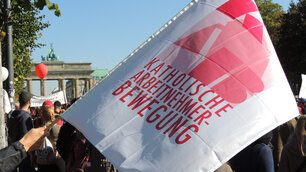 Eine weiße Fahne mit dem roten Aufdruck „KATHOLISCHE ARBEITNEHMER-BEWEGUNG“ wird bei einer Demonstration vor dem Brandenburger Tor in Berlin in die Höhe gehalten.