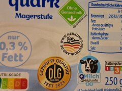 Die Nahaufnahme einer Quark-Verpackung zeigt verschiedene Gütesiegel und Nährwertangaben, darunter den Nutri-Score „A“, das DLG-Qualitätssiegel 2024, Hinweise auf die Haltungsform und die deutsche Herkunft sowie die Angabe „nur 0,3 % Fett“.